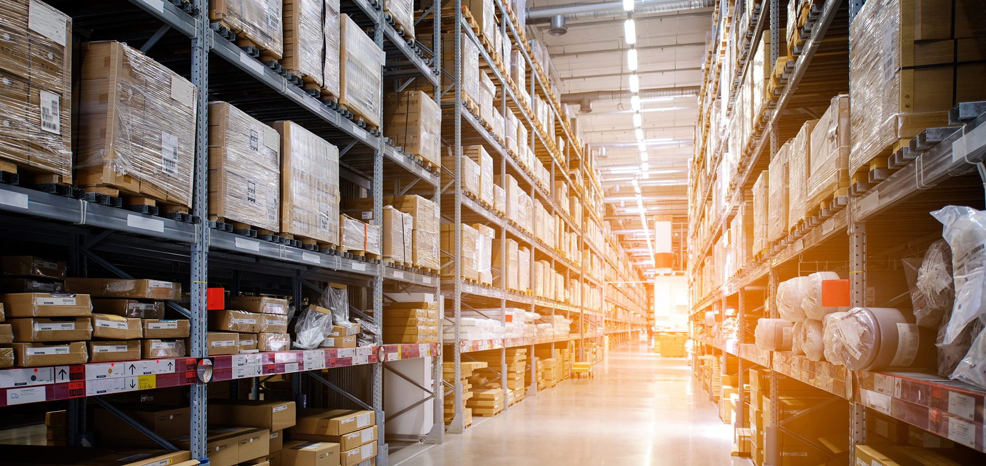 View down the glowing isle of a warehouse with boxes stacked on shelves to the ceiling